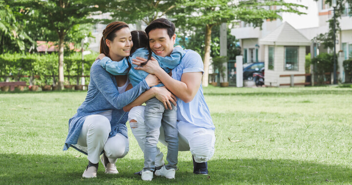 A mother and father hugging their daughter in a beautiful green garden near a housing area