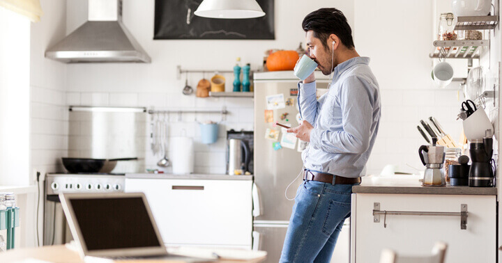 A man using his phone while drinking coffee alone at home with his computer ready for work.